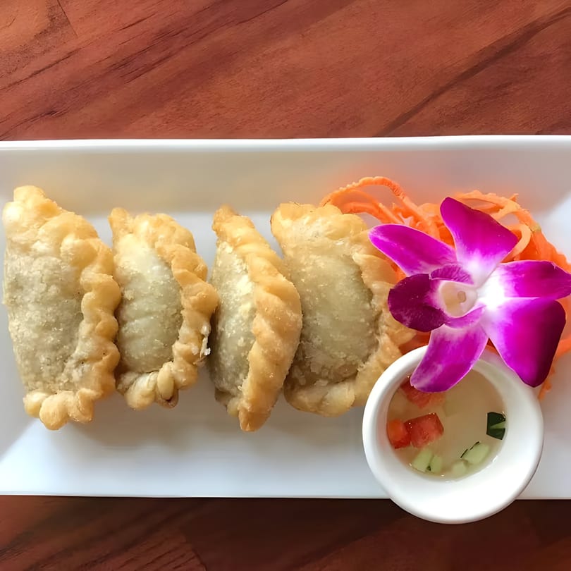 Four Thai curry puffs with a side of dipping sauce, shredded carrots, and a purple orchid.