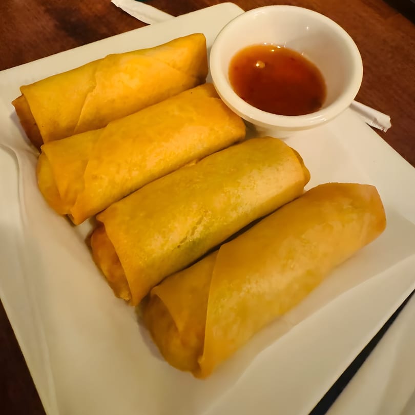 Four golden-brown Thai spring rolls and a small white bowl of dipping sauce on a white plate.